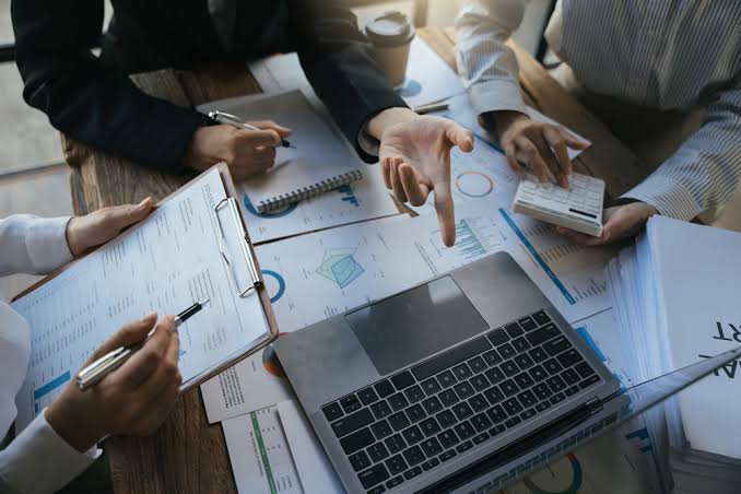 Vue rapprochée de mains au-dessus d'un bureau couvert de documents financiers et de graphiques, avec une calculatrice et un ordinateur portable.